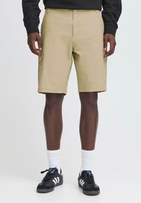 Beige cargo shorts med lige snit, bæltestropper, sidelommer og et glat bomuldsblandingsstof. Sammen med sorte sneakers og hvide sokker.