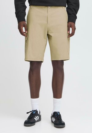 Pantalones cortos de carga beige con corte recto, lazos para cinturón, bolsillos laterales y una suave tela de mezcla de algodón. Combinados con zapatillas negras y calcetines blancos.