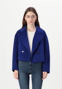 Kraftigt blå uldjakke med brede klape, dobbeltradet knaplukning og afslappet pasform, båret over en cremefarvet sweater og blå jeans.