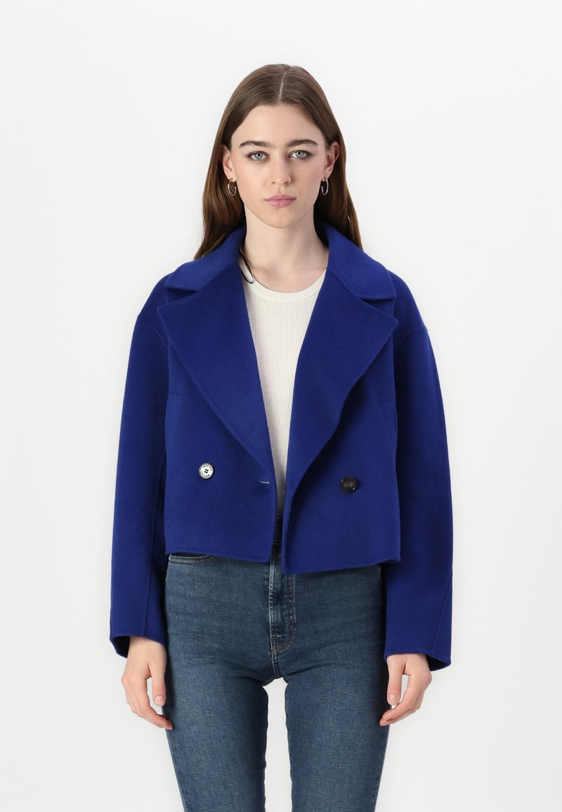 Kraftigt blå uldjakke med brede klape, dobbeltradet knaplukning og afslappet pasform, båret over en cremefarvet sweater og blå jeans.