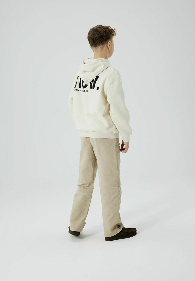 Felpa color crema con testo nero sul retro, abbinata a pantaloni beige e scarpe scure. Design semplice e texture morbida, adatta per un abbigliamento casual.