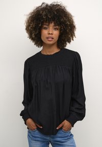 Frau mit lockigem Haar, die eine lockere schwarze Bluse mit gerafften Details und blaue Jeans trägt, steht mit den Händen in den Taschen vor einem schlichten Hintergrund.