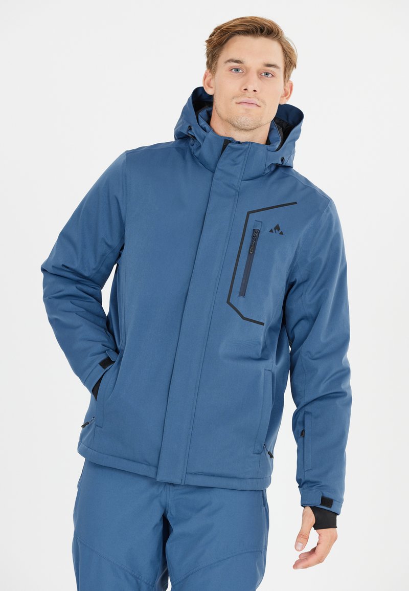 Junger Mann trägt eine blaue Winterjacke mit Kapuze und passende Hose, steht mit einer Hand in der Tasche vor weißem Hintergrund.