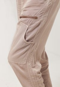 Gros plan sur un pantalon cargo marron clair montrant une poche zippée et une grande poche latérale ouverte sur la cuisse.