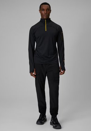 Męski model ubrany w czarny, długi top z suwakiem i otworami na kciuki, czarne spodnie cargo oraz czarne trampki, stojący na szarym tle.