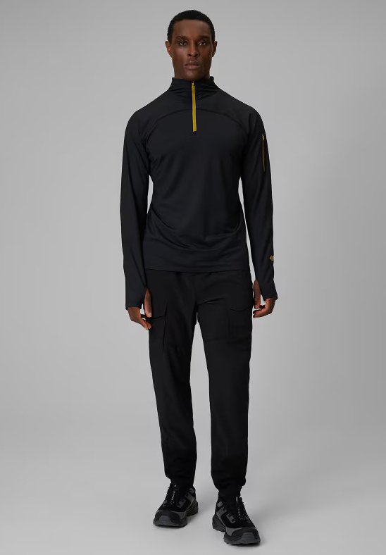 Mannequin homme portant un haut noir à manches longues zippé avec des trous pour les pouces, un pantalon cargo noir et des baskets noires, debout devant un fond gris.