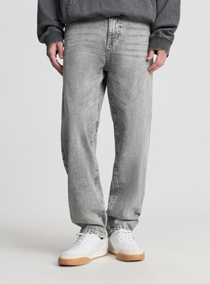 Osoba ubrana w wyblakłe szare dżinsy, białe trampki z gumową podeszwą oraz ciemnoszarą bluzę, stojąca na jednolitym tle.