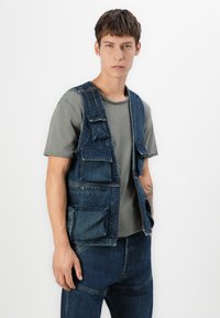 Νεαρός άνδρας που φοράει γκρι κοντομάνικο πουκάμισο, μπλε τζιν γιλέκο με πολλές τσέπες και αντίστοιχα μπλε τζιν παντελόνι, που στέκεται μπροστά από λευκό φόντο.