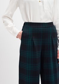 Hoch taillierte karierte Hose in Marineblau und Grün, mit Falten und seitlichen Taschen, kombiniert mit einer weißen Bluse mit langen Ärmeln.