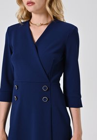 Königsblaue Wickelkleid aus glattem Stoff, mit Dreiviertelärmeln, goldenen Akzenten an den doppelreihigen Knöpfen und einer taillierten Silhouette.