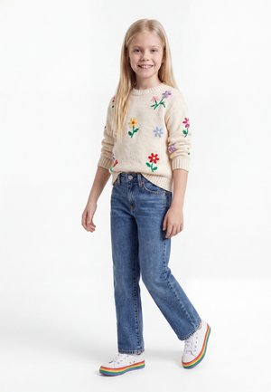 Κορίτσι με μακριά ξανθά μαλλιά στέκεται χαμογελαστή, φοράει μια κρεμ πουλόβερ με κεντημένα λουλούδια, μπλε τζιν και λευκά αθλητικά με πολύχρωμες σόλες.