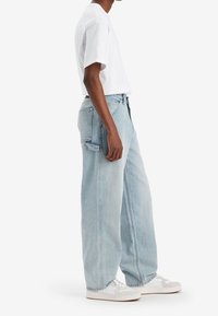 Jeans de mezclilla de color azul claro y pernera ancha; cuenta con un diseño clásico de cinco bolsillos y dobladillo recto. Combinado con una camiseta de manga corta blanca.