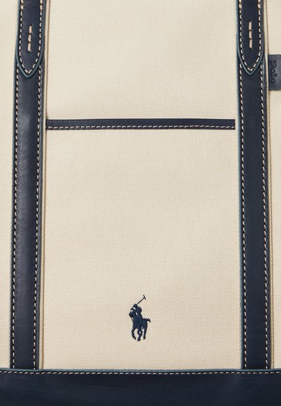 Beige stof med navy blå læderstropper og syninger langs kanterne, med en lille navy blå broderet polo spiller logo.
