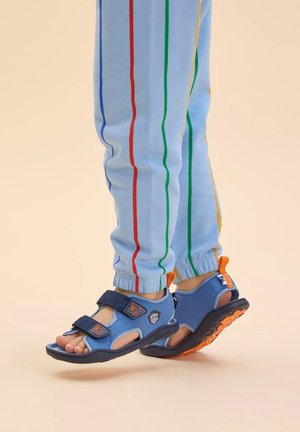 Blaue Sportsandalen mit verstellbaren Klettverschlüssen; verfügen über Fußausschnitte, orangefarbene Akzente und eine strukturierte schwarze Sohle. Kombiniert mit hellblauen gestreiften Hosen.