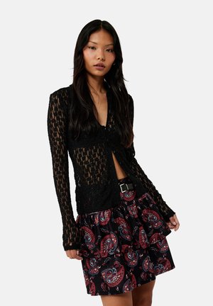 Jonge vrouw met lang donker haar die een zwarte kanten top met lange mouwen en een zwarte rok met een rood paisleymotief en gelaagde ruches draagt.