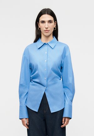 Γυναίκα με μακριά σκούρα μαλλιά που φορά χρυσούς κρίκους στα αυτιά, μπλε πουκάμισο με κουμπιά και λευκές ρίγες, και σκούρο παντελόνι, στέκεται μπροστά σε λευκό φόντο.