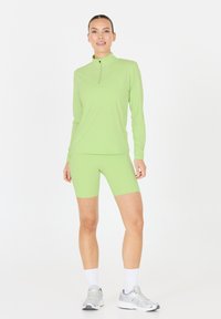 Femme debout portant un haut zippé à manches longues vert clair et un short de vélo assorti, avec des chaussettes blanches et des baskets argentées sur fond blanc.