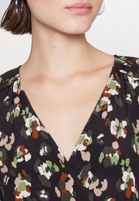 Czarna kopertowa bluzka w kwiatowy wzór z zielonymi, pomarańczowymi i kremowymi akcentami; lekka tkanina z dekoltem w kształcie litery V i marszczonymi ramionami.
