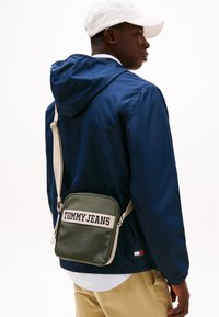 Borsa a tracolla verde Tommy Jeans con forma rettangolare, tracolla beige e logo in testo bianco. Indossata sopra una giacca blu navy e pantaloni color khaki.