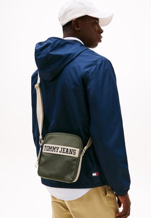 Grønn Tommy Jeans crossover-veske med rektangulær form, beige stropp og hvit tekstlogo. Bært over en marineblå jakke og kaki bukser.