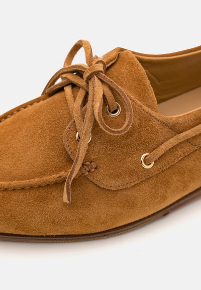 Mocassino in suede marrone con occhielli dorati, lacci annodati e un design cucito lungo il bordo. Suola liscia.