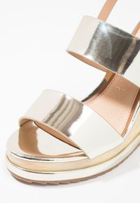 Sandale à plateforme argentée métallique avec deux larges brides, un bout arrondi et une semelle intérieure beige texturée. La semelle extérieure est en caoutchouc beige pour une meilleure adhérence.