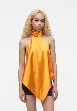 Jeune femme aux longs cheveux blonds bouclés portant un haut orange brillant à col montant et ourlet asymétrique sur un short noir.