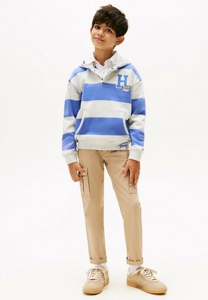 Niño de pie vestidos con una sudadera de rayas azules y grises, camisa de cuello blanco, pantalones cargo beige y zapatillas beige sobre un fondo blanco.