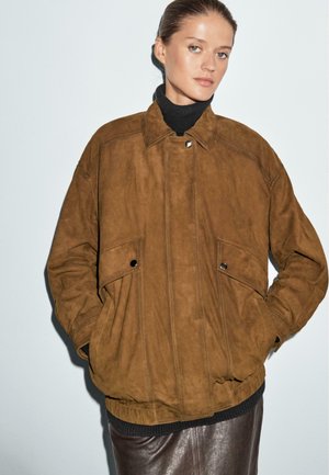 Woman wearing a brown suede jacket with flap pockets over a black turtleneck and dark brown leather skirt, standing against a white wall.