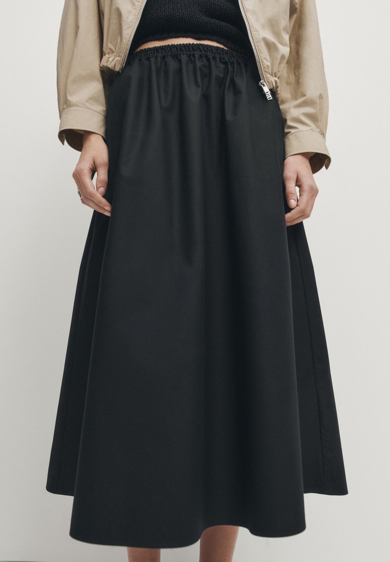 Femme portant une jupe midi noire à taille élastique, un haut noir et une veste courte beige, debout devant un fond clair uni.
