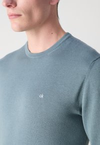 Homme portant un pull léger bleu clair à col rond avec un petit logo blanc "ck" sur la poitrine, visible des épaules au menton.