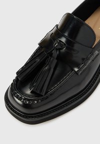Crne kožne loafers cipele s sjajnom završnom obradom, koje imaju dekorativne resice, detalje šivanja i debeli crni potplat.