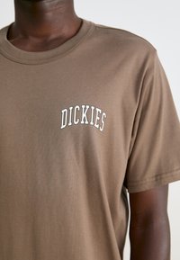Camiseta de algodón marrón con cuello redondo y un logo blanco en forma de arco "DICKIES" en el lado izquierdo del pecho. Textura suave, ajuste relajado.