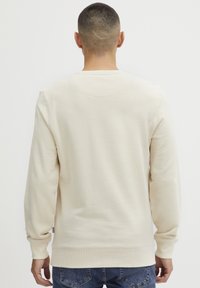 Sweatshirt couleur crème à manches longues, avec des poignets côtelés et un col rond. Texture douce, design minimaliste. Porté avec un jean bleu.