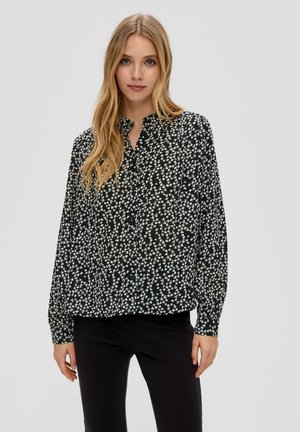 Zwarte blouse met een wit bloemenpatroon, zonder kraag, lange mouwen, vloeiende stof en lichtjes geraffineerde zoom. Draagt bij zwarte broek.