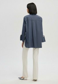 Navy blue oversized shirt with a high neckline, long sleeves, and side slits. Paired with cream-colored pants and silver-accented shoes.