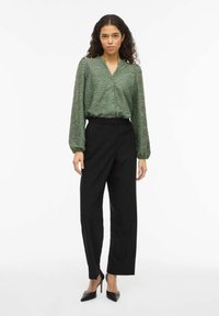 Blusa verde con estampado, escote en V y cierre de botones, combinada con pantalones negros de tiro alto y tacones negros puntiagudos.