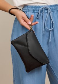 Schwarze Leder-Clutch mit gefaltetem Design, die eine glatte Textur und einen bronzefarbenen Verschluss zeigt. In einer Hand gehalten, die sich in der Nähe von hellblauen Hosen befindet.
