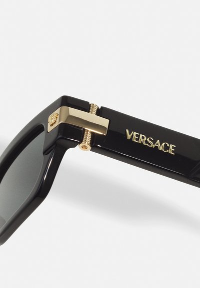 Zwarte zonnebril met een glossy afwerking, voorzien van gouden accenten en het "VERSACE" logo aan de zijkant, geometrisch frameontwerp en donkere lenzen.