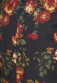 Svart blomsterblus med mönster av röda, gula och gröna blommor, beige knappar och en texturerad tyg med en lätt glansig yta.