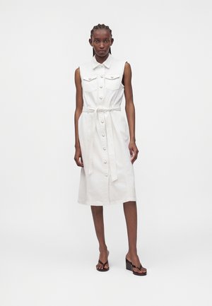 Γυναίκα που φοράει λευκό μίντι φόρεμα χωρίς μανίκια με κουμπιά, γιακά, τσέπες στο στήθος, ζώνη στη μέση και μαύρα τακούνια σανδάλια, που στέκεται μπροστά σε λευκό φόντο.