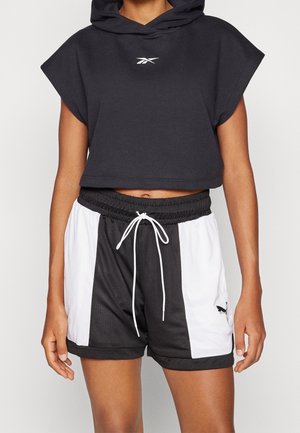 Persoon draagt een zwarte cropped mouwloze hoodie en zwart-witte sportshorts met trekkoord en Puma-logo op het linkerbeen.