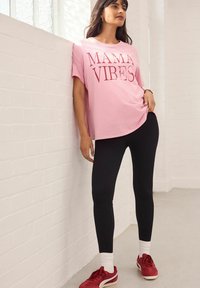 Lichtroze katoenen t-shirt met "MAMA VIBES" in donkere tinten gedrukt, gecombineerd met zwarte geribde leggings en rode sneakers met witte accenten.