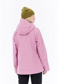 Blonde person wearing a green beanie and pink hooded jacket, standing facing away on a white background.