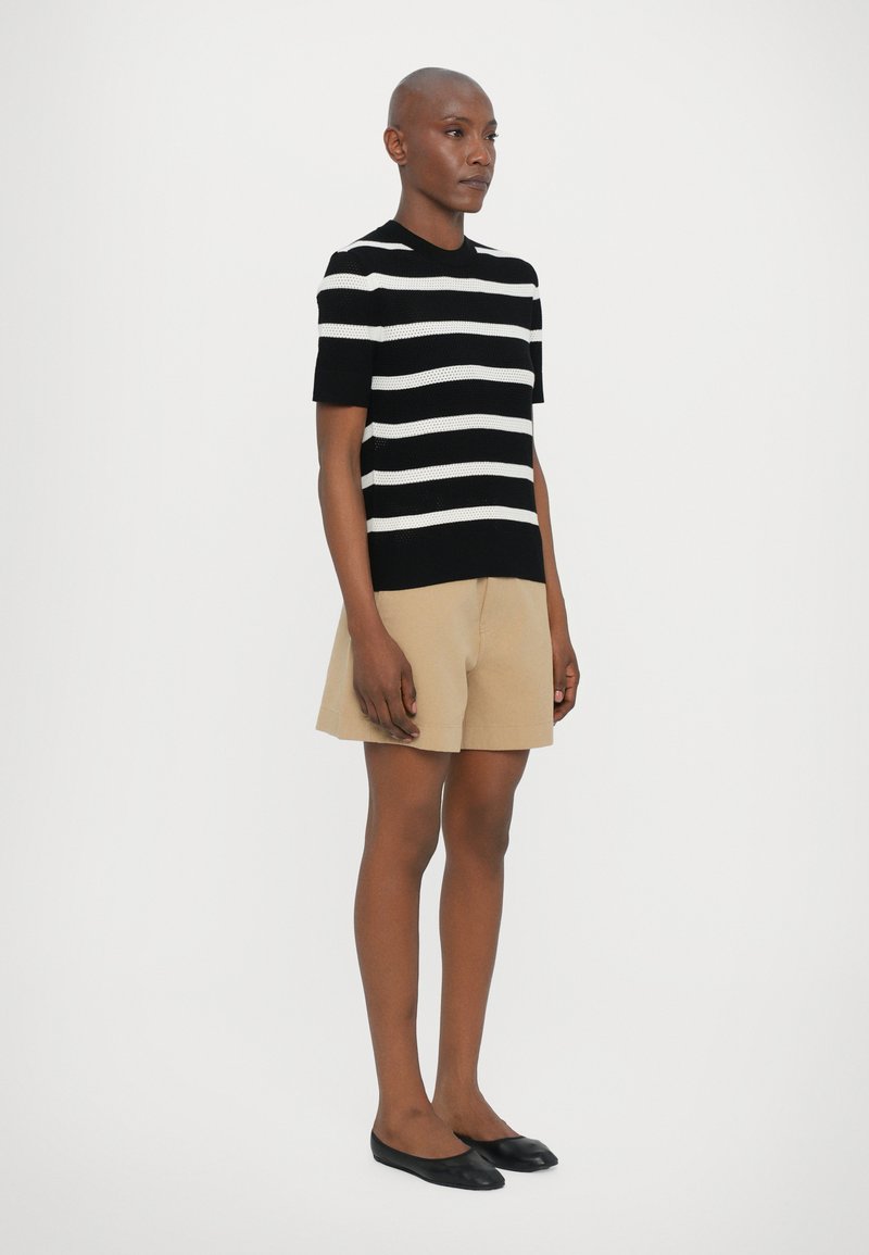 Haut en tricot rayé noir et blanc à manches courtes, associé à un short beige. Le mannequin porte des chaussures plates noires et se tient devant un fond blanc.