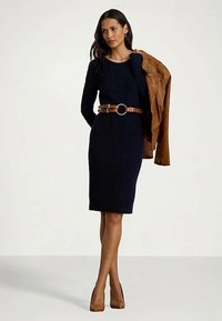 Moteris, vilkinti tamsiai mėlyną suknelę su rudonu diržu, laiko rudą odinę striukę per vieną pečių, avėdama rudus aukštakulnius batus, stovinti ant vientiso fono.