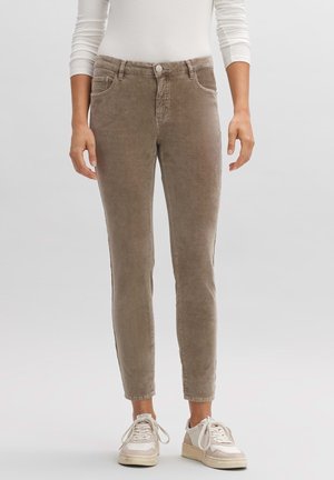 Osoba ubrana w brązowe dopasowane spodnie sztruksowe i białe trampki, stojąca na tle gładkiego, jasnoszarego tła.