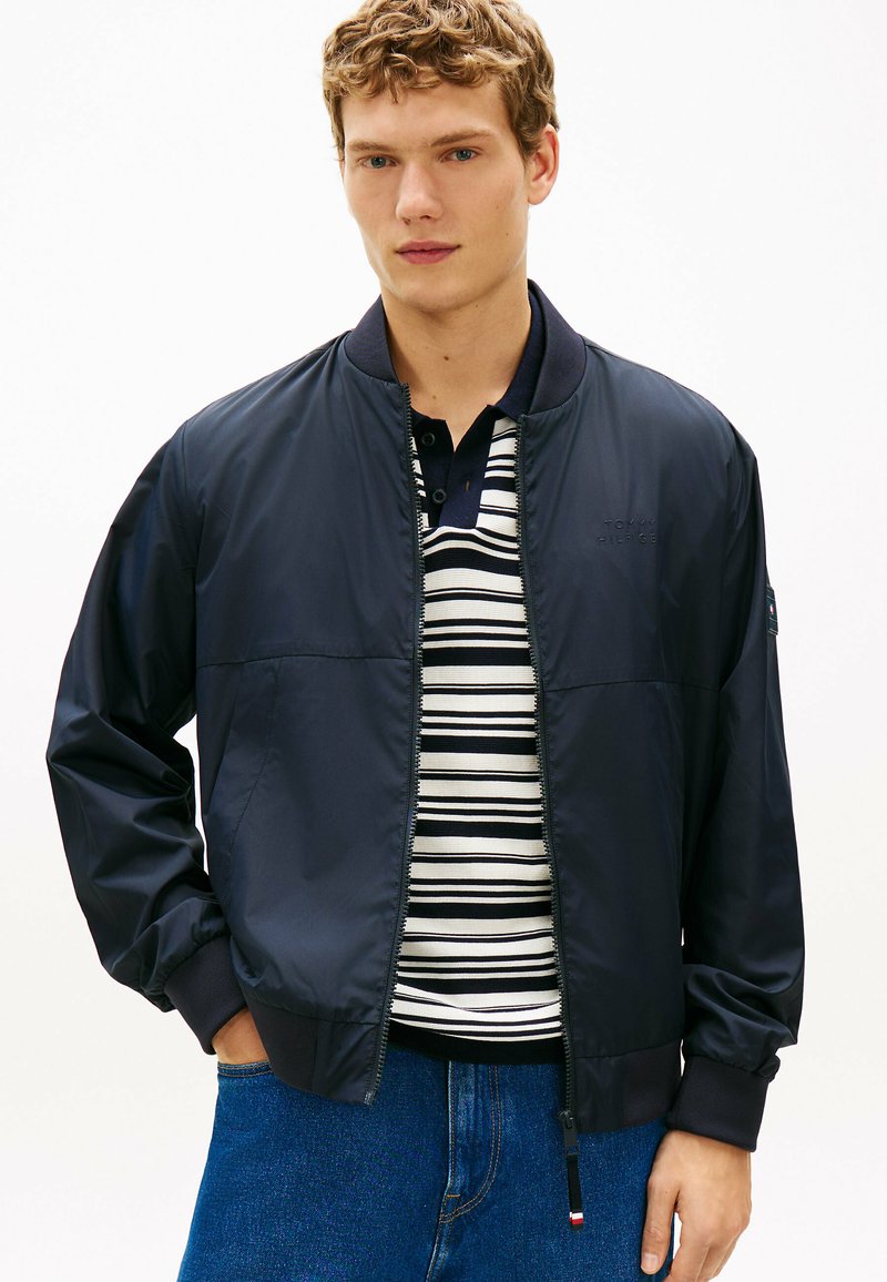 Jeune homme portant une veste bomber bleu marine sur un polo à rayures noires et blanches et un jean bleu, posant devant un fond blanc.