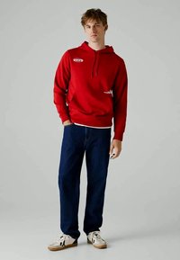 Jovem de pé, vestindo um hooded vermelho, jeans azul escuro e ténis brancos com detalhes pretos, contra um fundo cinza liso.