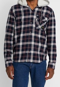Plaid flannel shirt in navy, red, and white, featuring a soft grey-lined hood, button-down front, and a chest pocket with patch details.